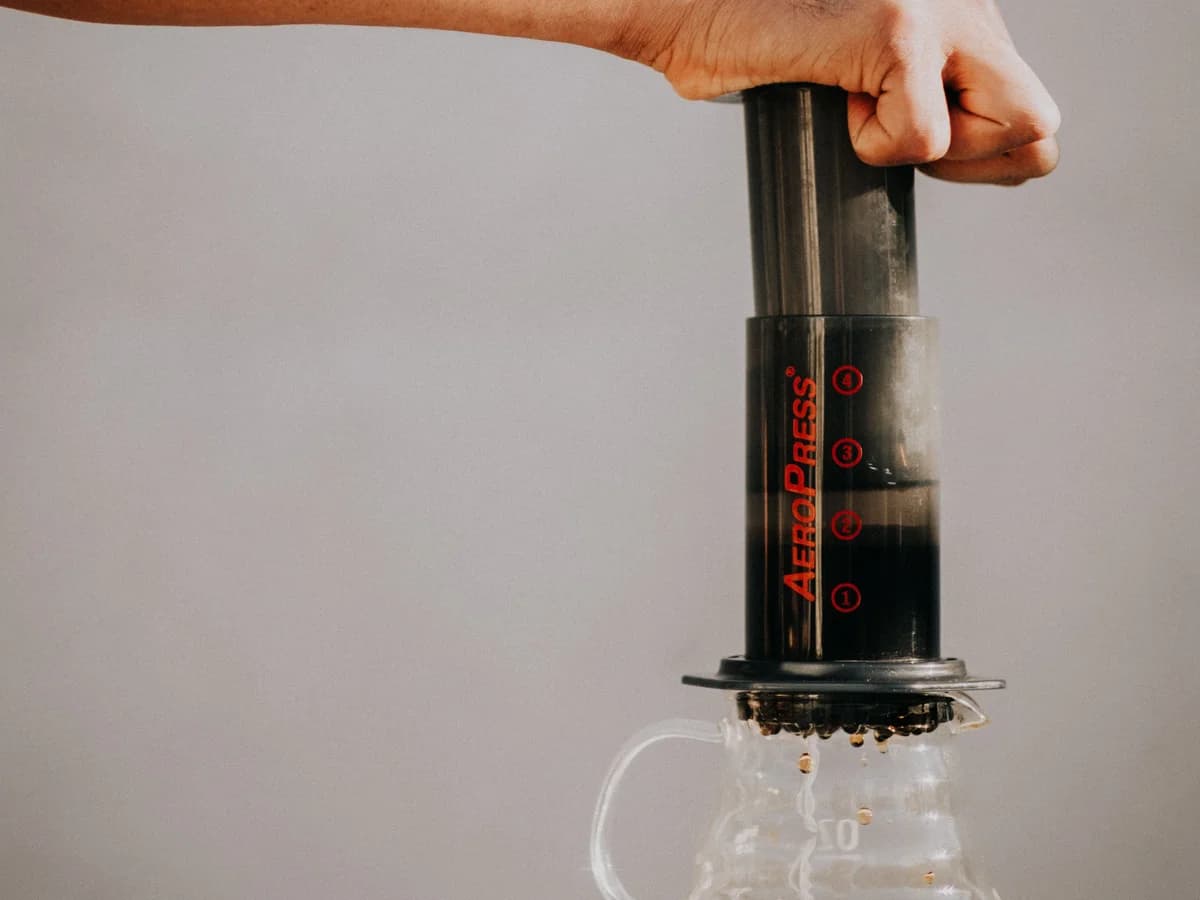 A hand pressing the plunger of an AeroPress mounted on a glass carafe.