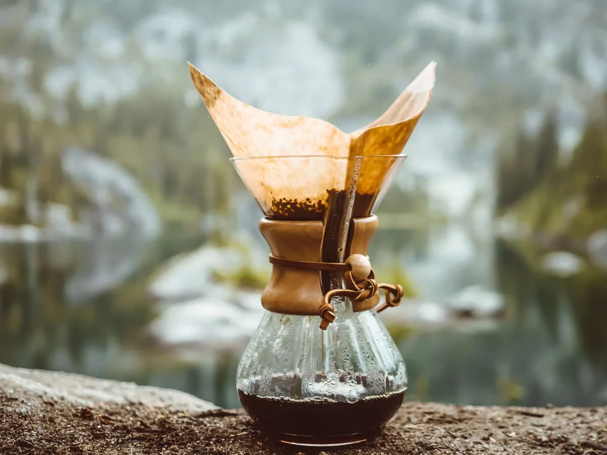 An hourglass-shaped Chemex with a folded paper filter and wooden collar, brewed coffee in the base, set outdoors.