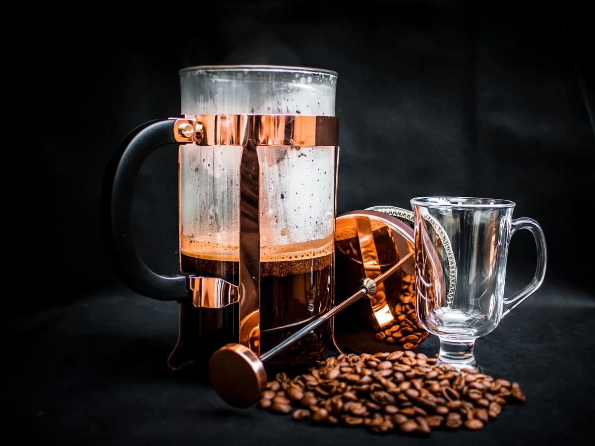 A glass French press part-filled with brewed coffee, its plunger lifted out, beside scattered beans and a glass mug.