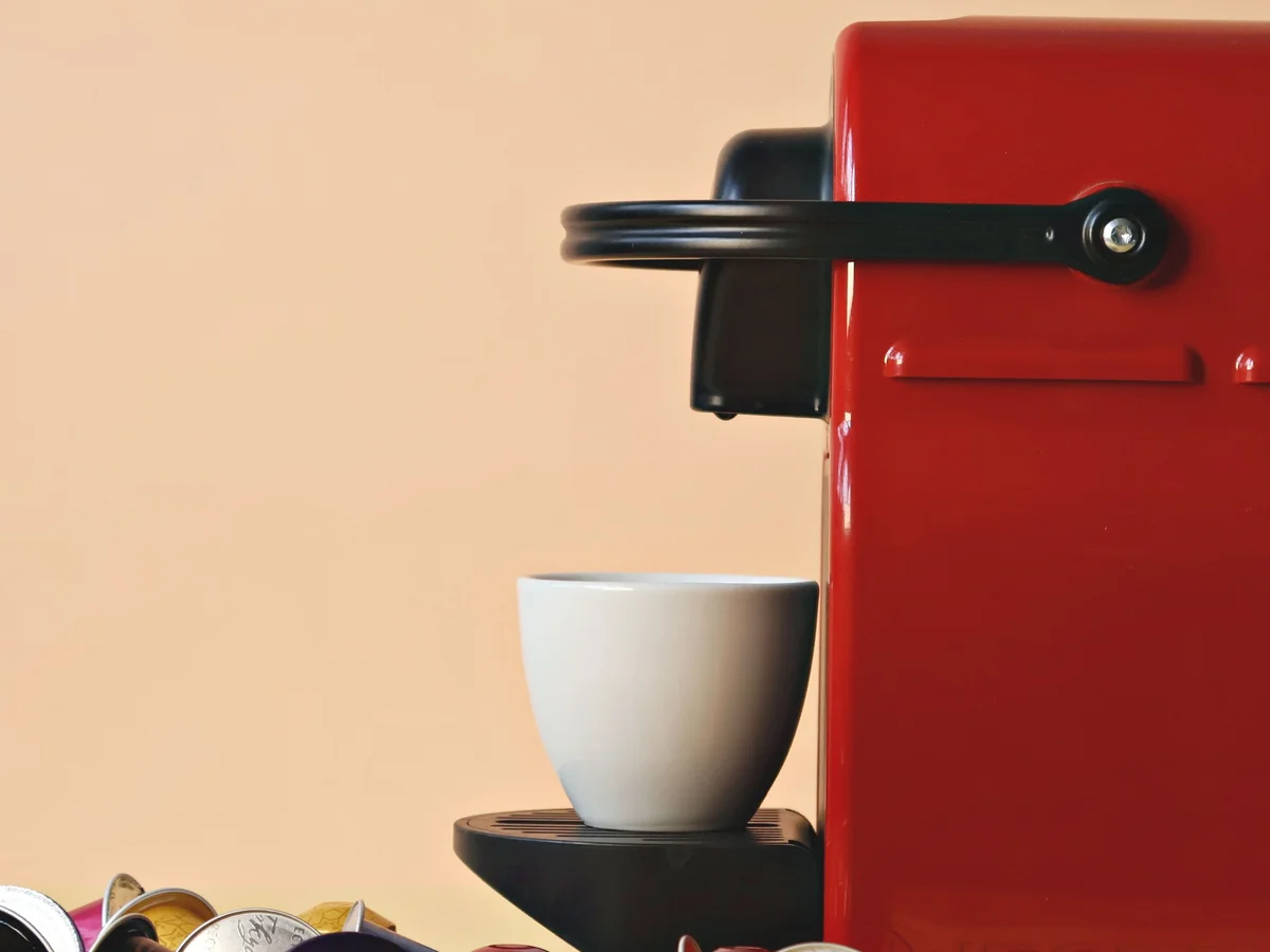 A red capsule coffee machine with its lever closed and a white cup on the drip tray, coloured capsules in front.