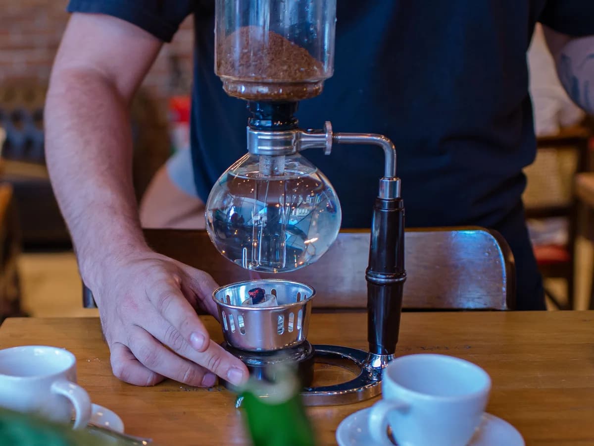 A person tending a siphon brewer, coffee grounds in the upper glass chamber and water in the lower globe over a burner.