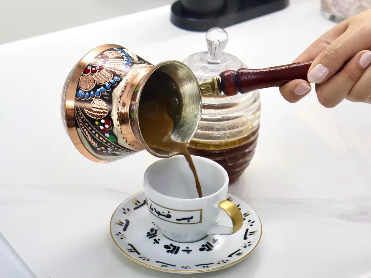 A decorated copper cezve tipped to pour Turkish coffee into a small patterned cup and saucer.