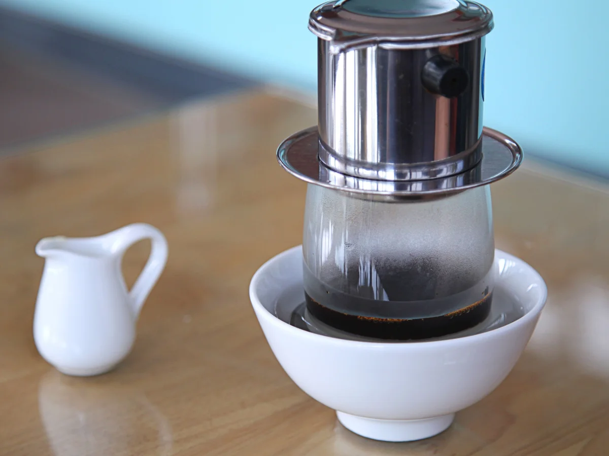 A stainless steel phin filter resting on a glass set in a white bowl, brewed coffee collecting below.