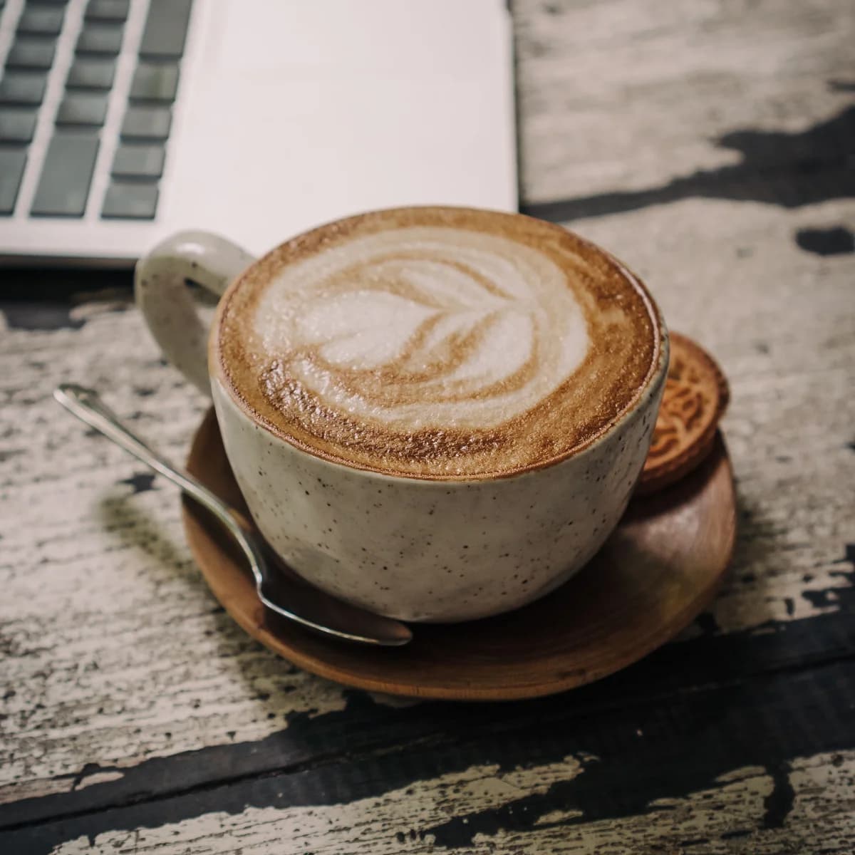 A speckled ceramic cup of cappuccino with a thick foam layer and a leaf pattern on top, on a wooden saucer.