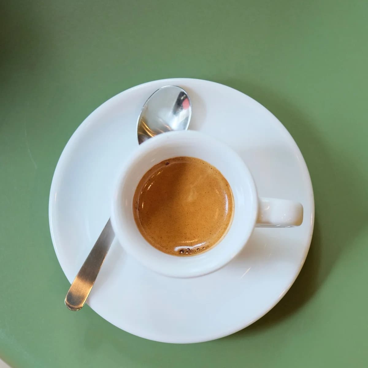 A small white cup of espresso topped with hazelnut-coloured crema, on a saucer with two spoons.