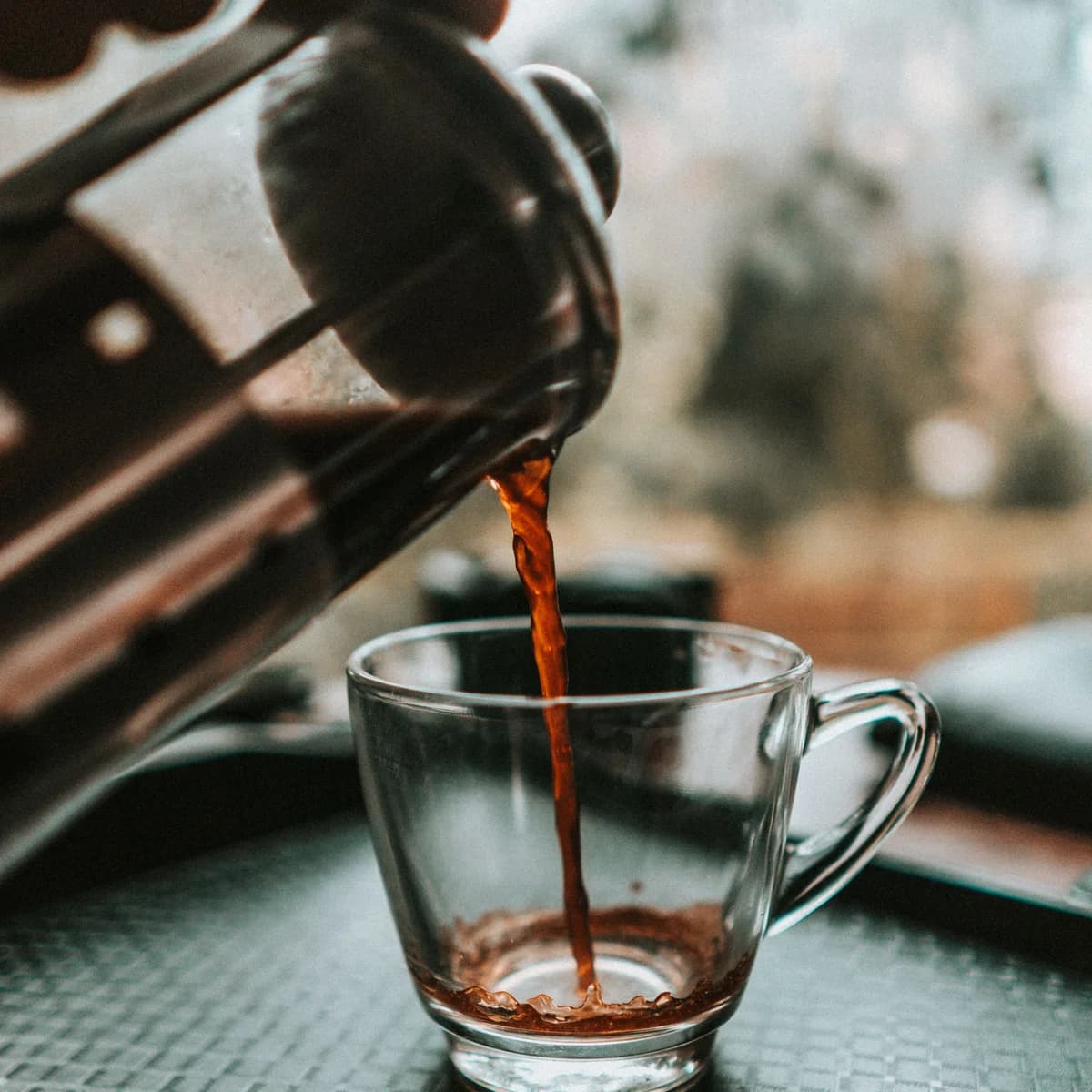 Dark brewed coffee being poured from a French press into a clear glass cup.