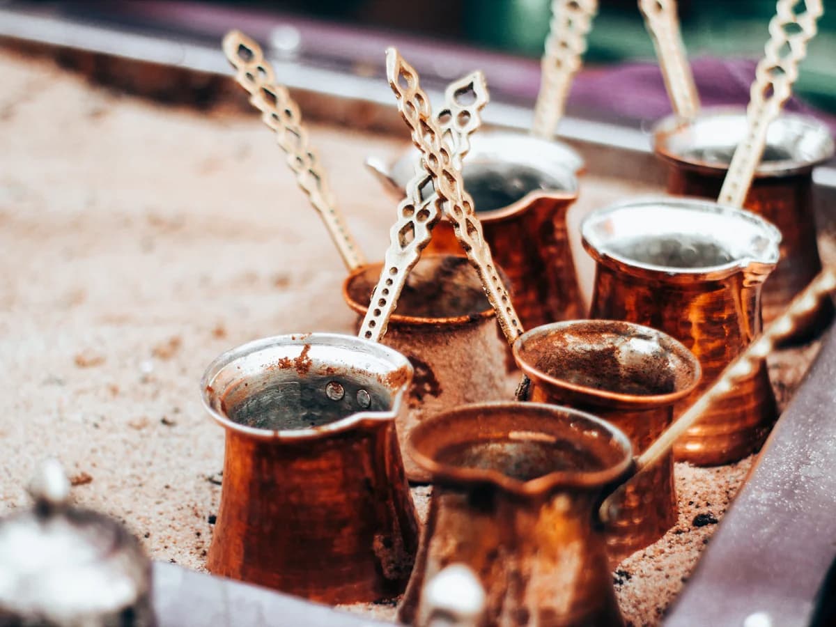 Several hammered copper cezves with long, pierced metal handles standing in a tray of hot sand, some part-filled with coffee.