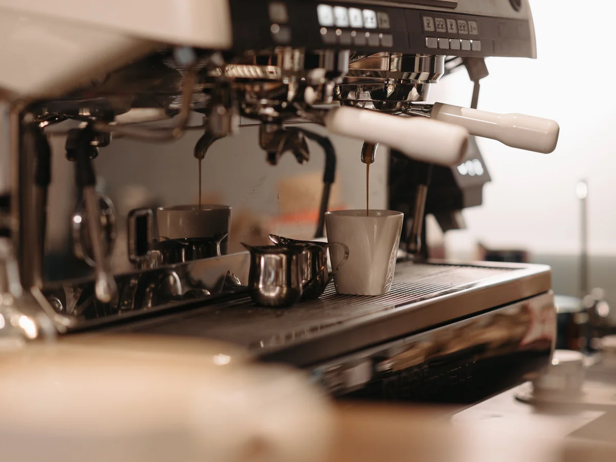Stainless group heads on an espresso machine with pale-handled portafilters locked in, two dark streams of espresso running into cups on the drip tray.