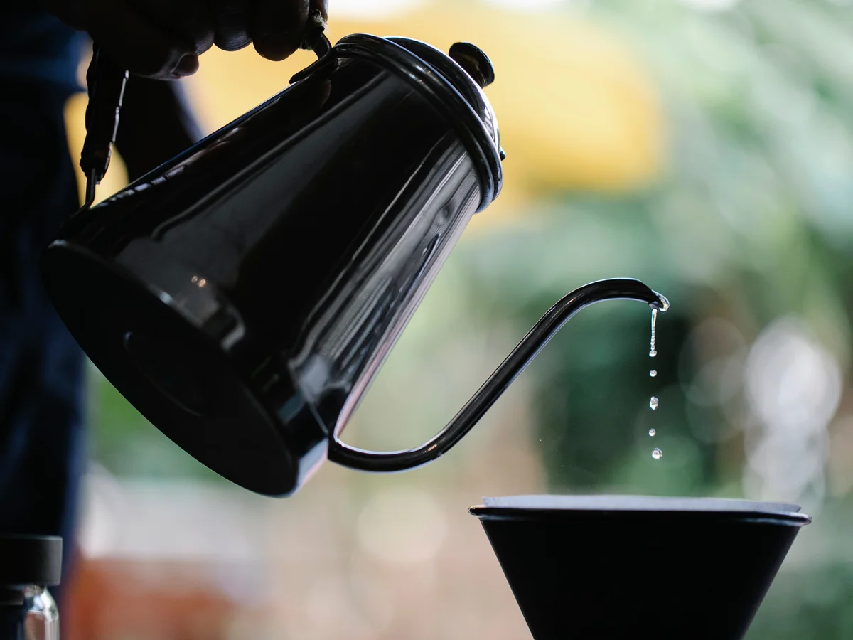A dark gooseneck kettle tipped forward in profile, its narrow S-curved spout releasing a thin line of water droplets into a black cone dripper below.