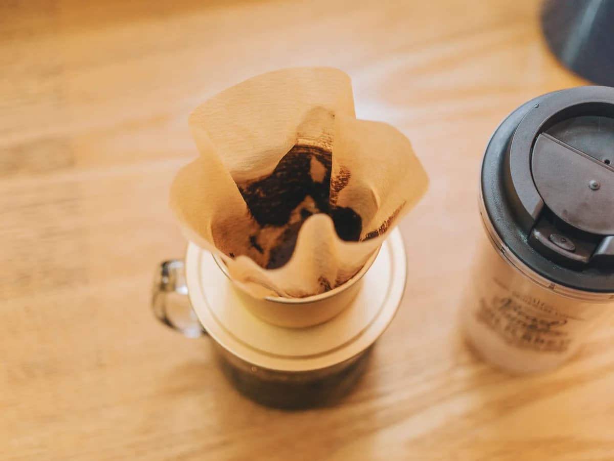 A fluted brown paper coffee filter holding spent grounds, seated in a white cone dripper resting on a glass mug of brewed coffee.