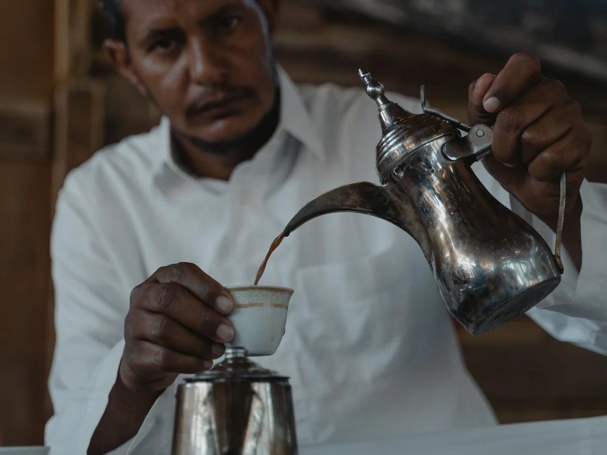 A man in a white shirt pouring Arabic coffee from a long-spouted dallah into a small handleless cup.