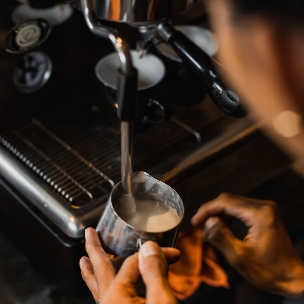 A barista steaming milk in a stainless steel jug held under an espresso machine's steam wand.