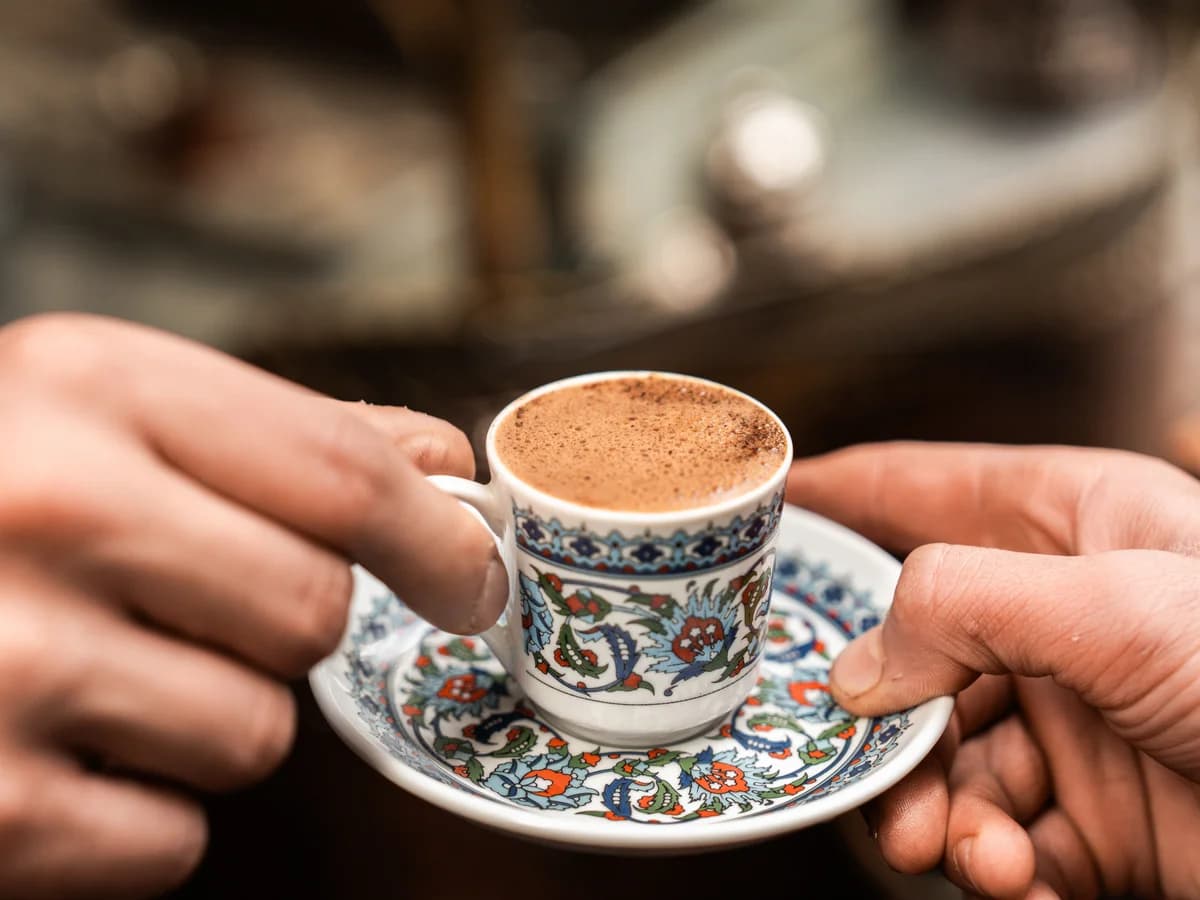 Hands passing a small patterned cup of Turkish coffee on a matching saucer.